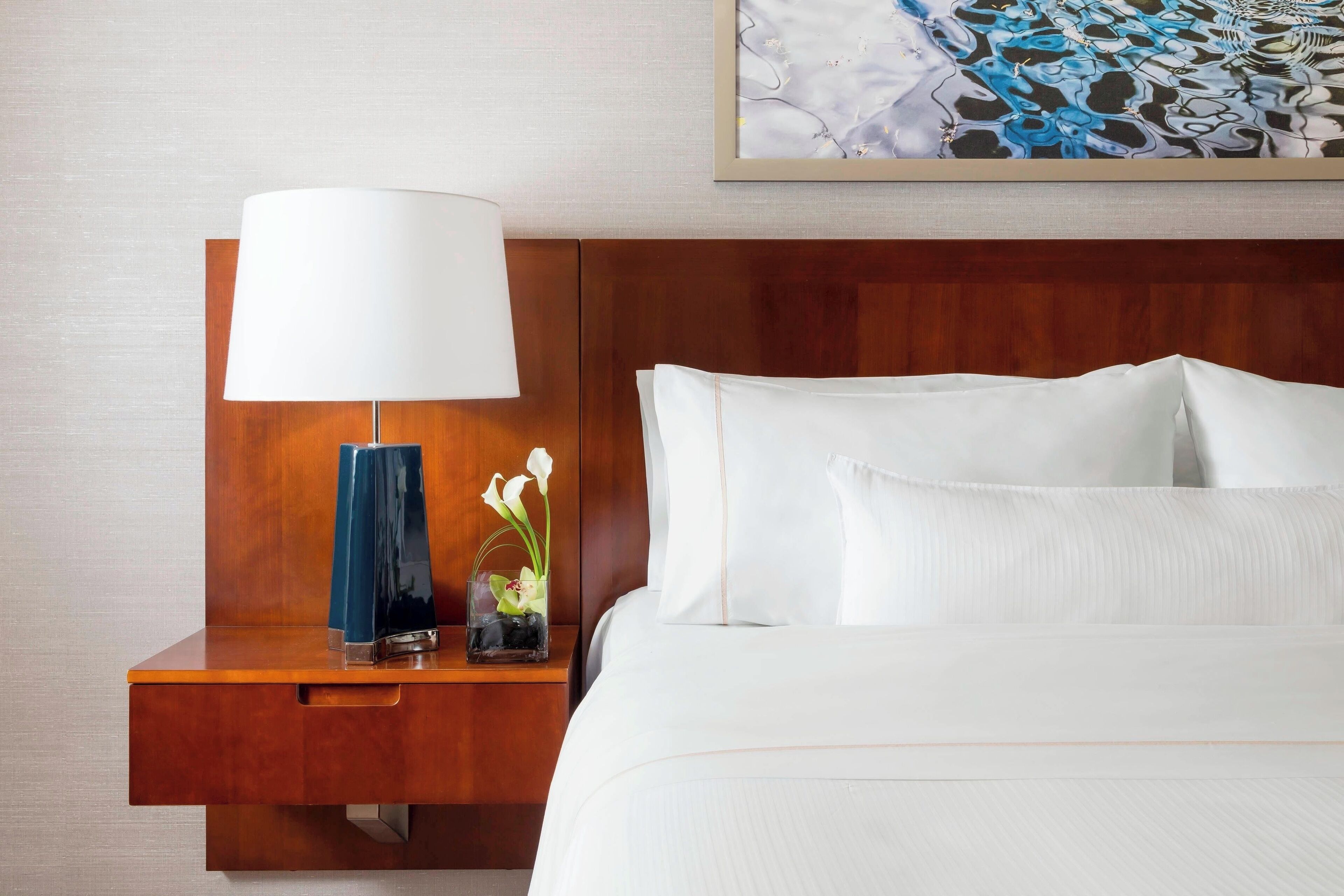 A hotel room with a wooden headboard, a bedside table with a lamp, a vase with flowers, and a bed with white linens.