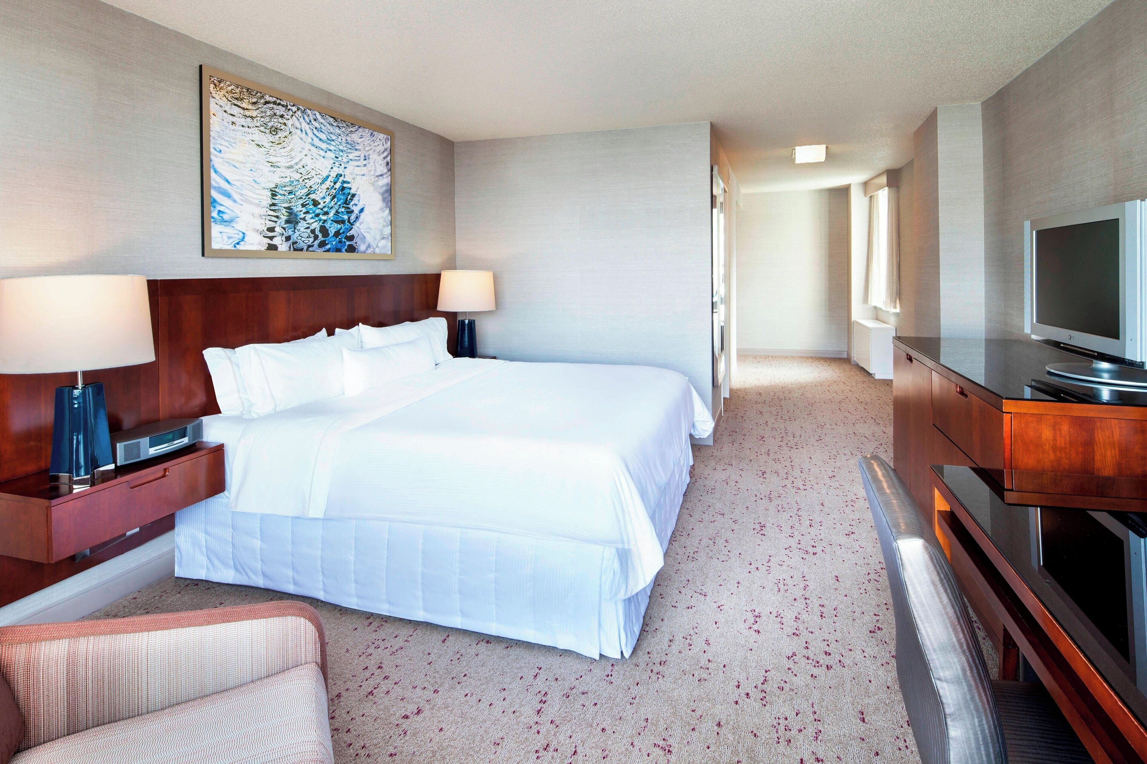 A hotel room with a large bed, a desk with a lamp, a television, and a painting on the wall.