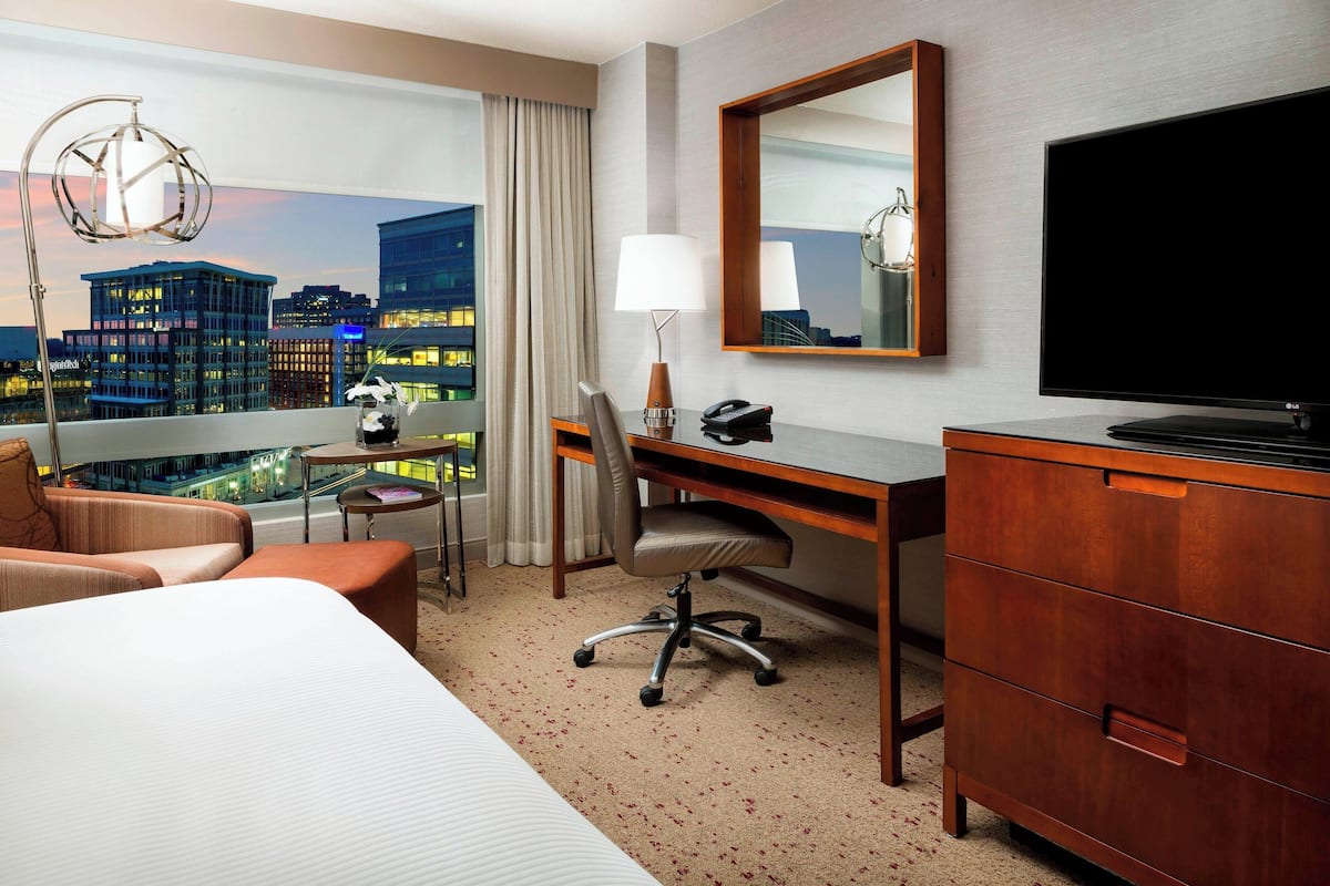 Quarto de hotel com cama, escrivaninha, cadeira e cômoda. Vê-se a cidade pela janela, e há um abajur e uma TV de tela plana.