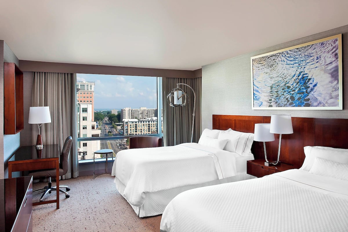 Quarto de hotel com duas camas, uma escrivaninha, uma cadeira, mesa de cabeceira com abajures e uma grande janela com vista para a cidade.