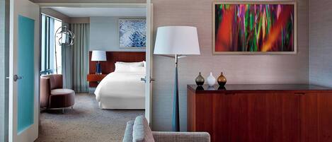 A hotel room with a bed, a desk, a chair, a lamp, and a painting on the wall.