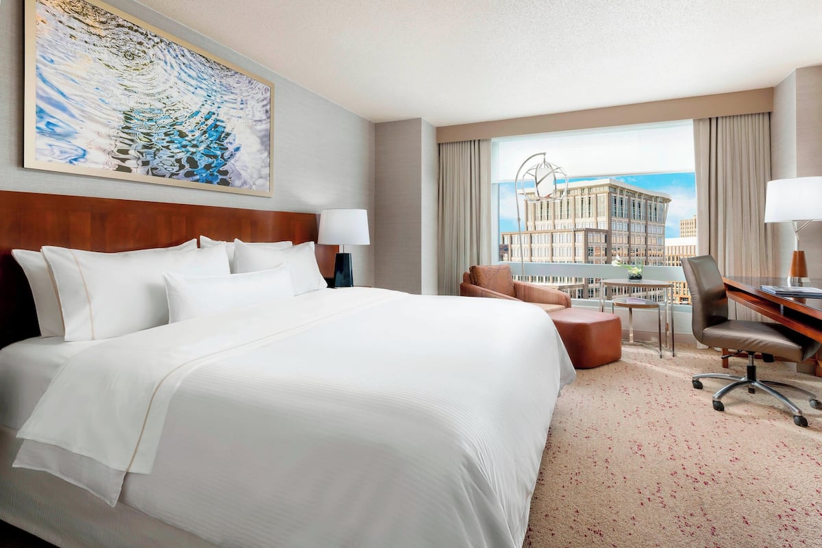 Quarto de hotel com uma cama grande, uma escrivaninha, uma cadeira e vista para um edifício pela janela.