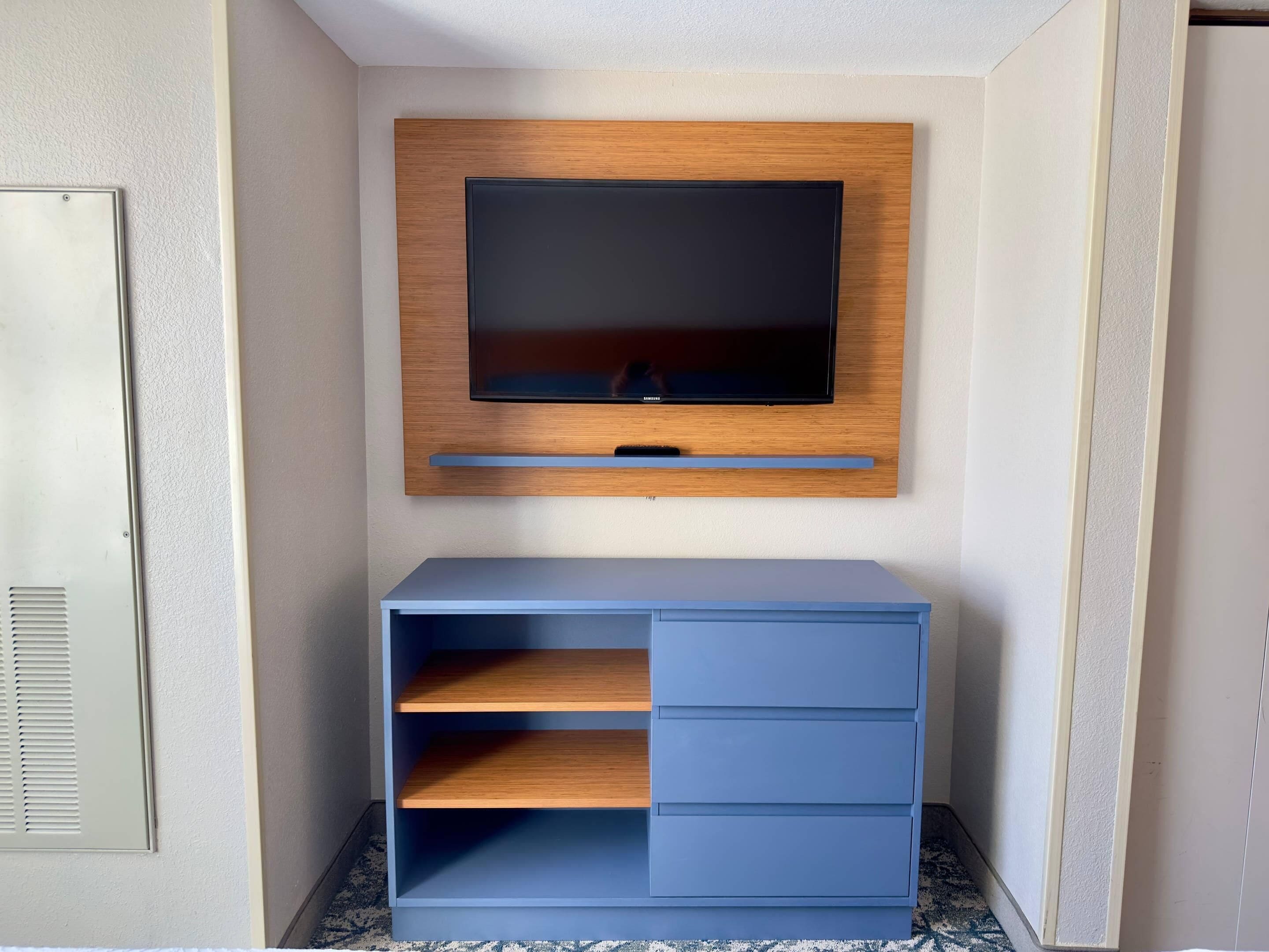 A modern room with a mounted TV on a wooden panel, a blue cabinet with drawers, and a vent on the left wall.
