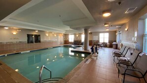 An indoor swimming pool with a diving board, tiled walls, and a seating area with chairs.