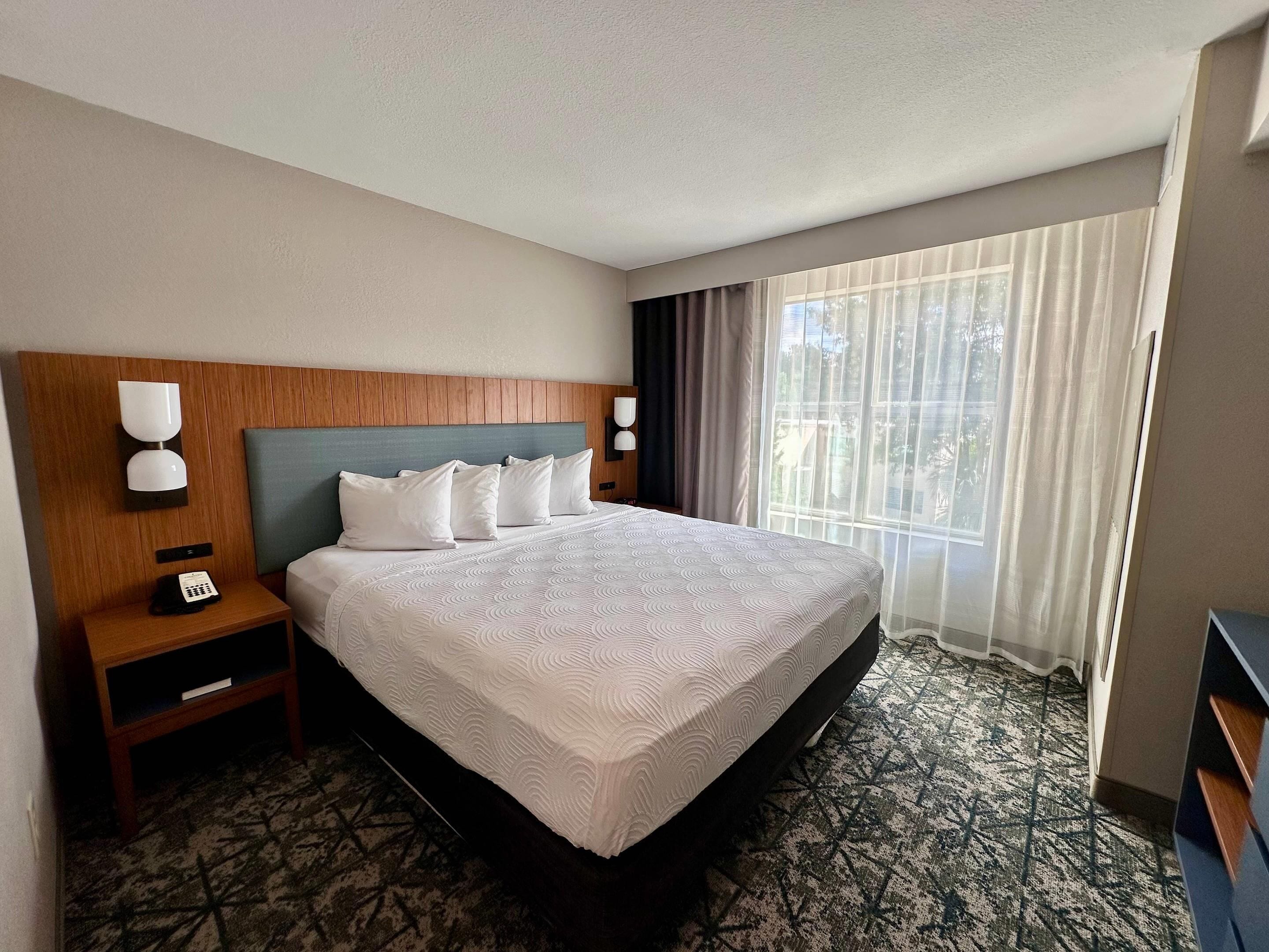 A hotel room with a large bed, a wooden headboard, two bedside tables with lamps, a telephone, and a window with curtains.