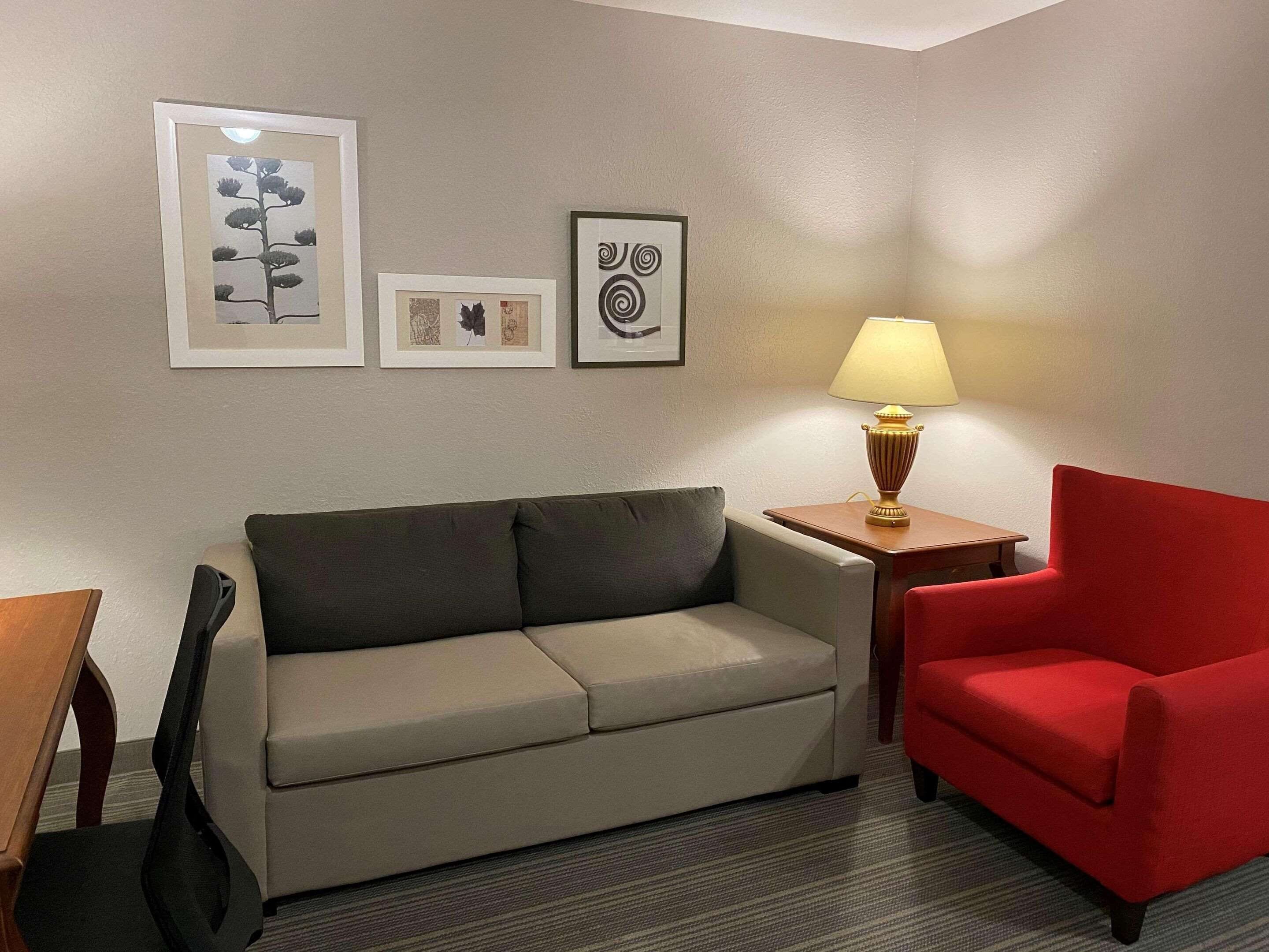 Ein Zimmer mit einem beigen Sofa, einem roten Sessel und einem kleinen Tisch mit einer Lampe. An der Wand hängen drei gerahmte Bilder.
