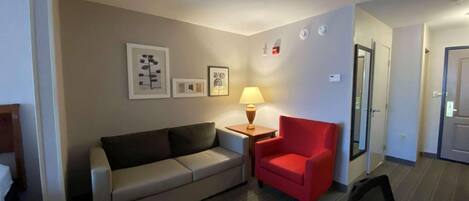 Une chambre d’hôtel avec un canapé, un fauteuil rouge, une petite table avec une lampe et des tableaux encadrés au mur.