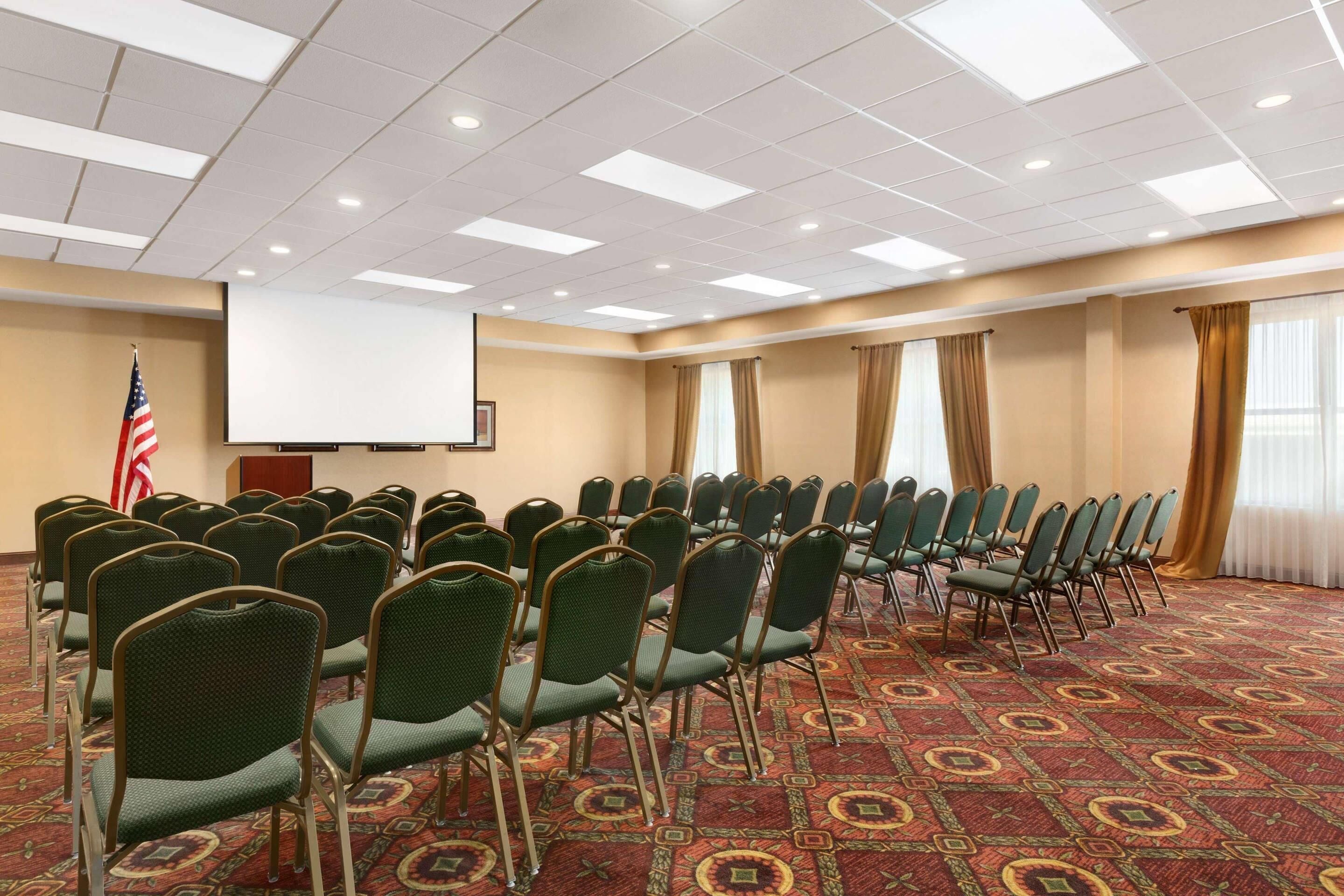 A conference room with rows of green chairs, a projector screen, an American flag, and a carpeted floor.