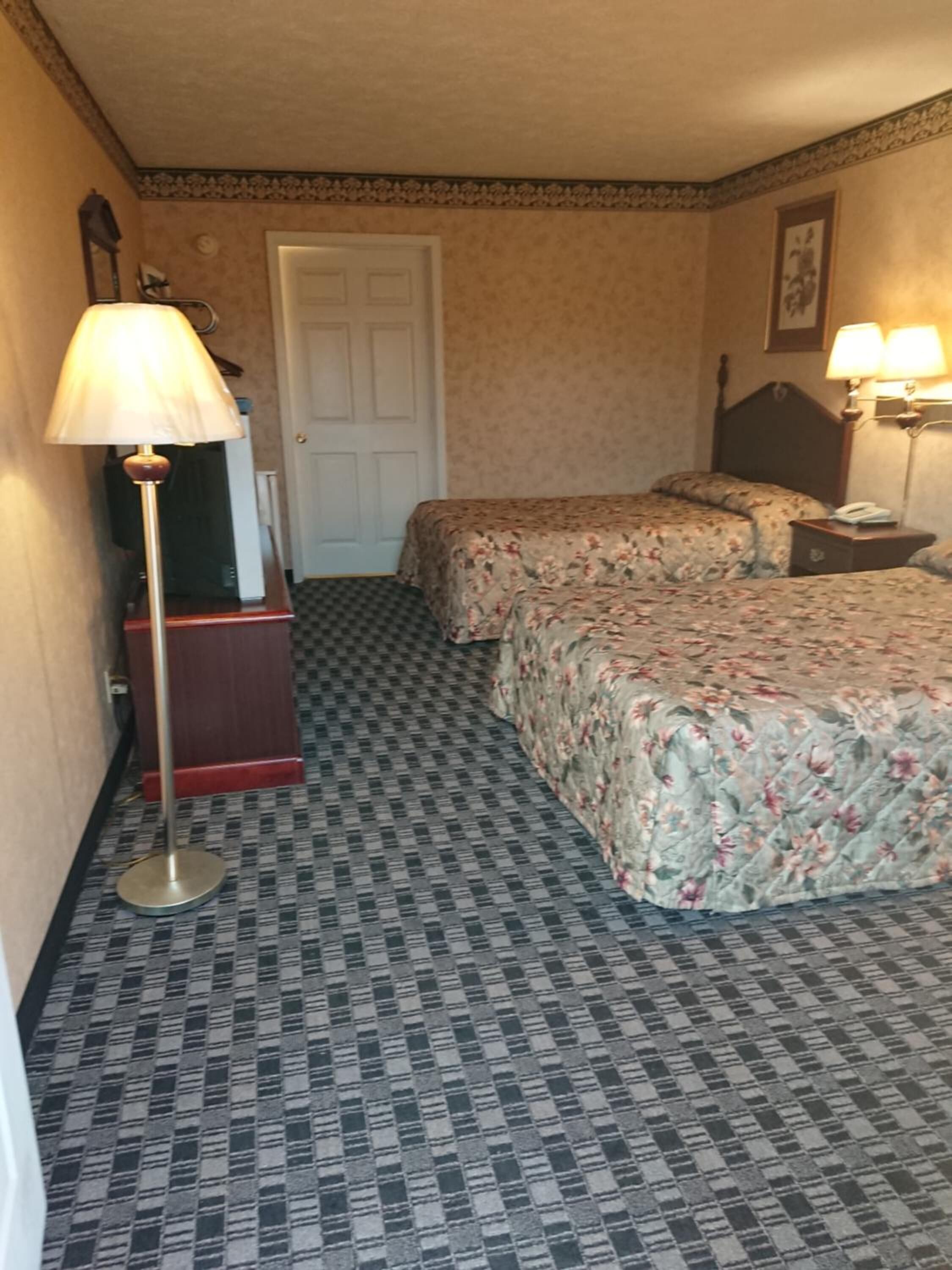 A hotel room with a checkered floor, two beds with floral bedspreads, a standing lamp, a door, and a framed picture on the wall.