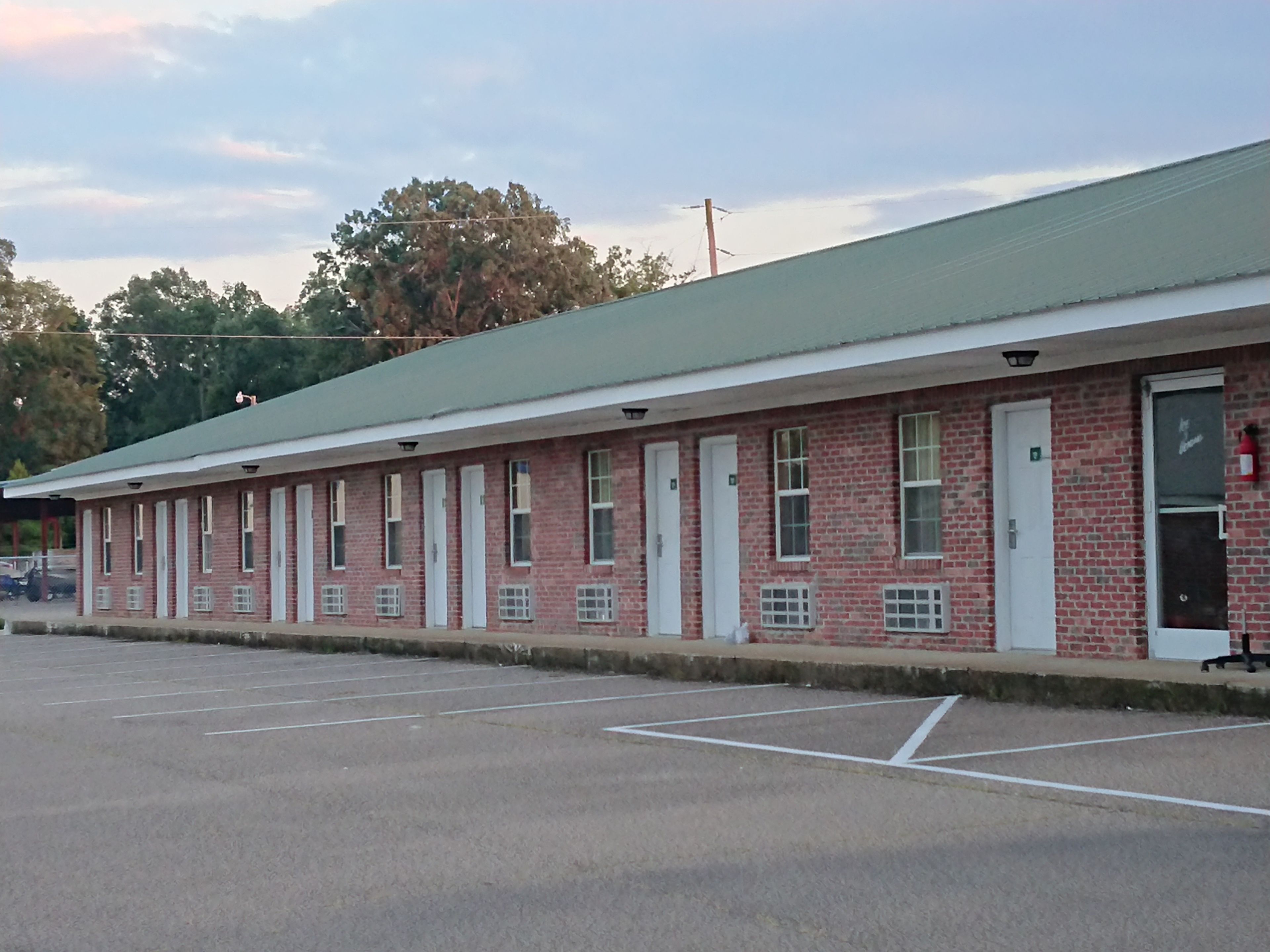 A row of single-story brick motel rooms with a green roof, a parking lot in front, and a tree in the background.