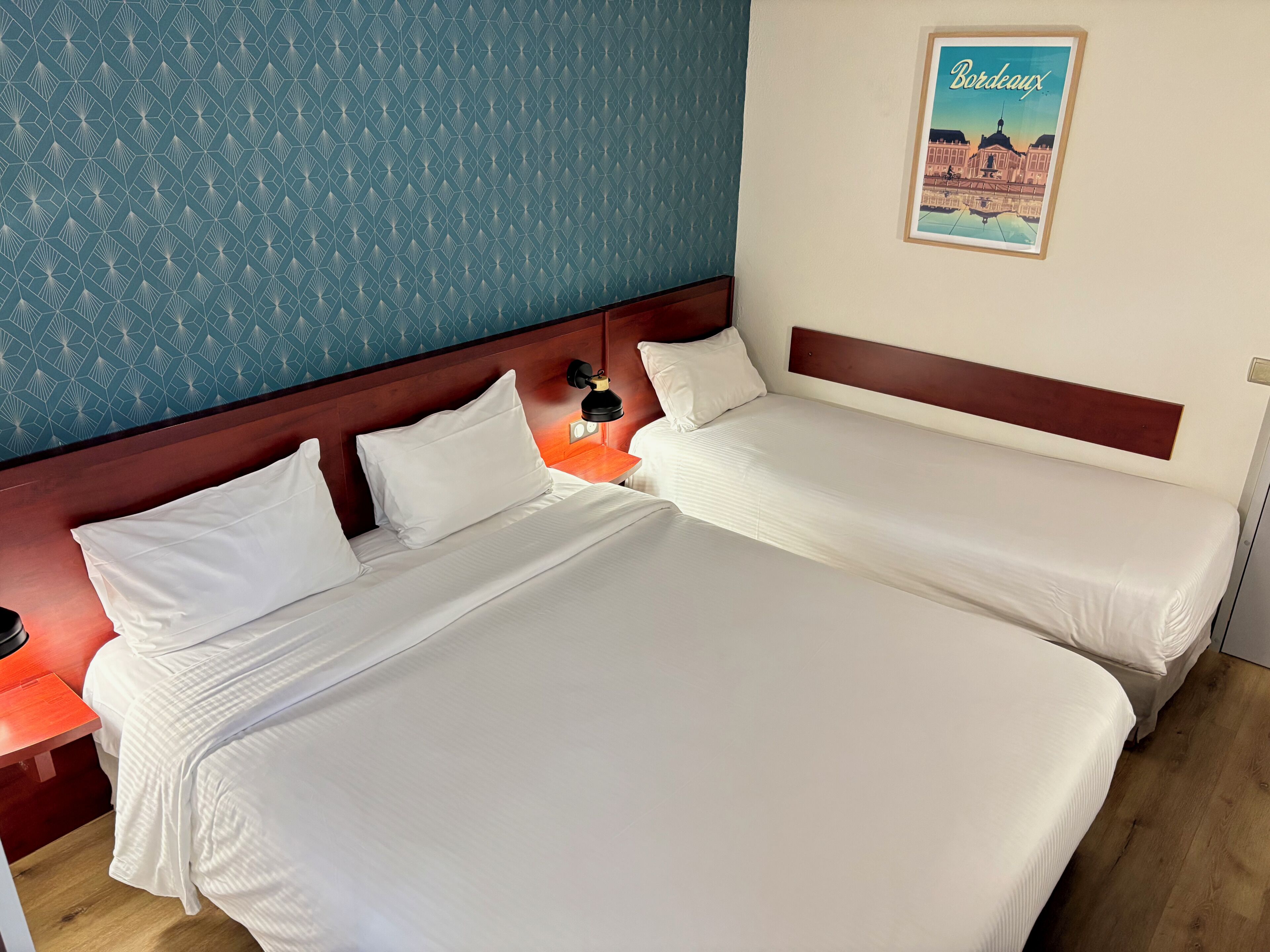 A hotel room with two beds, a nightstand with a lamp, a poster of Bordeaux, and a blue patterned wall.