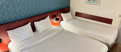 A hotel room with two beds, a nightstand with a lamp, a poster of Bordeaux, and a blue patterned wall.