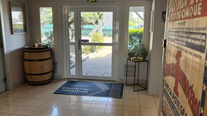 L’entrée d’un hôtel, avec un tonneau en bois, un vase en verre posé sur un piédestal et un paillasson.