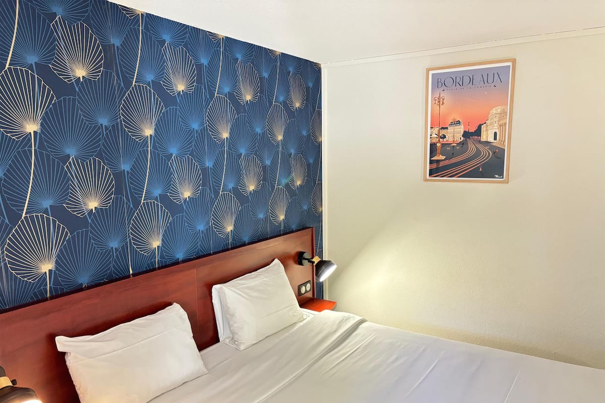 Quarto de hotel com cama, roupa de cama branca, cabeceira, mesa de cabeceira com abajur, parede com padrão de folhas azuis e um pôster de Bordeaux.