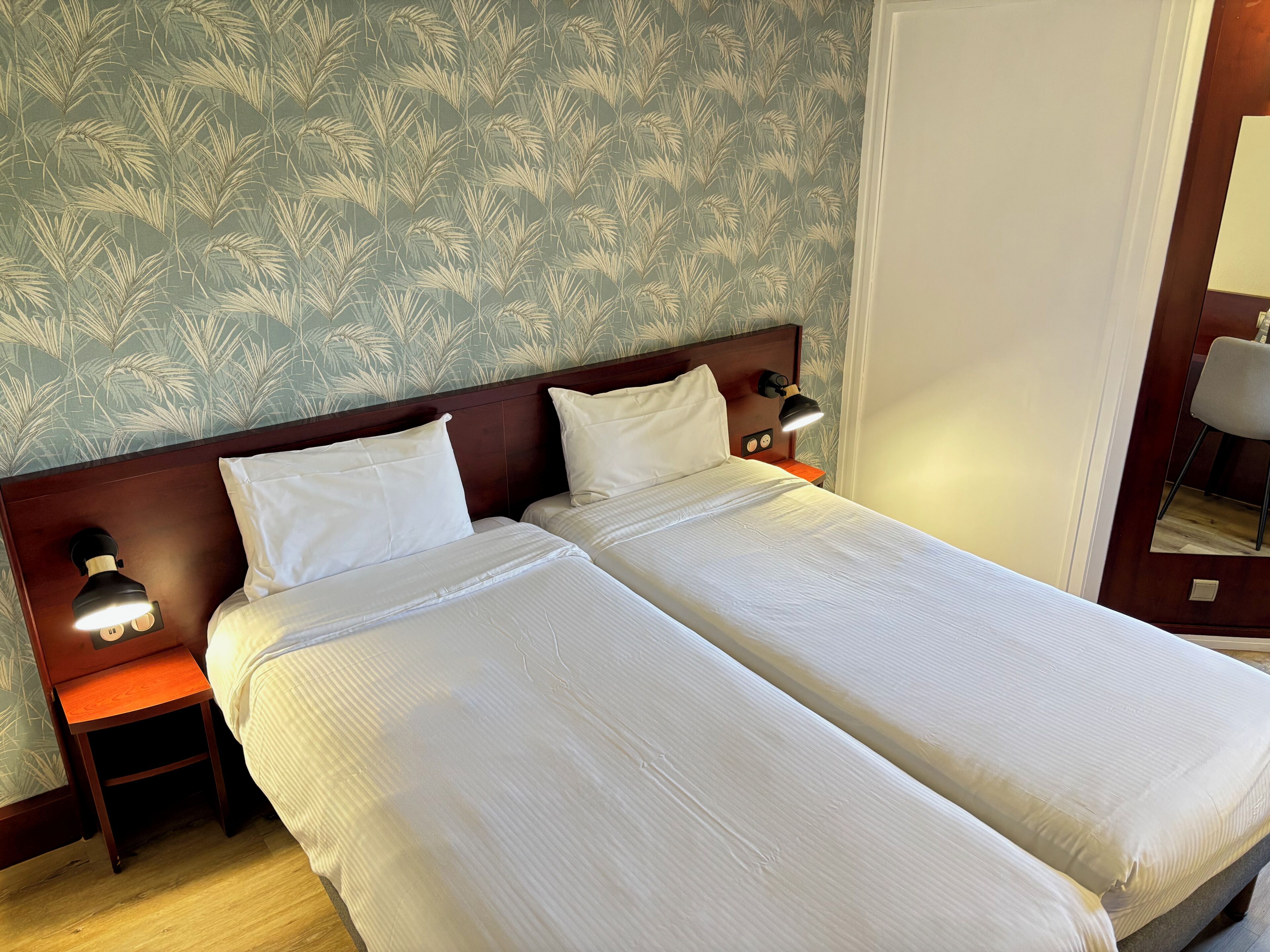 Una cama doble con sábanas blancas, dos mesitas de noche con lámparas, y un estampado floral en el papel tapiz.