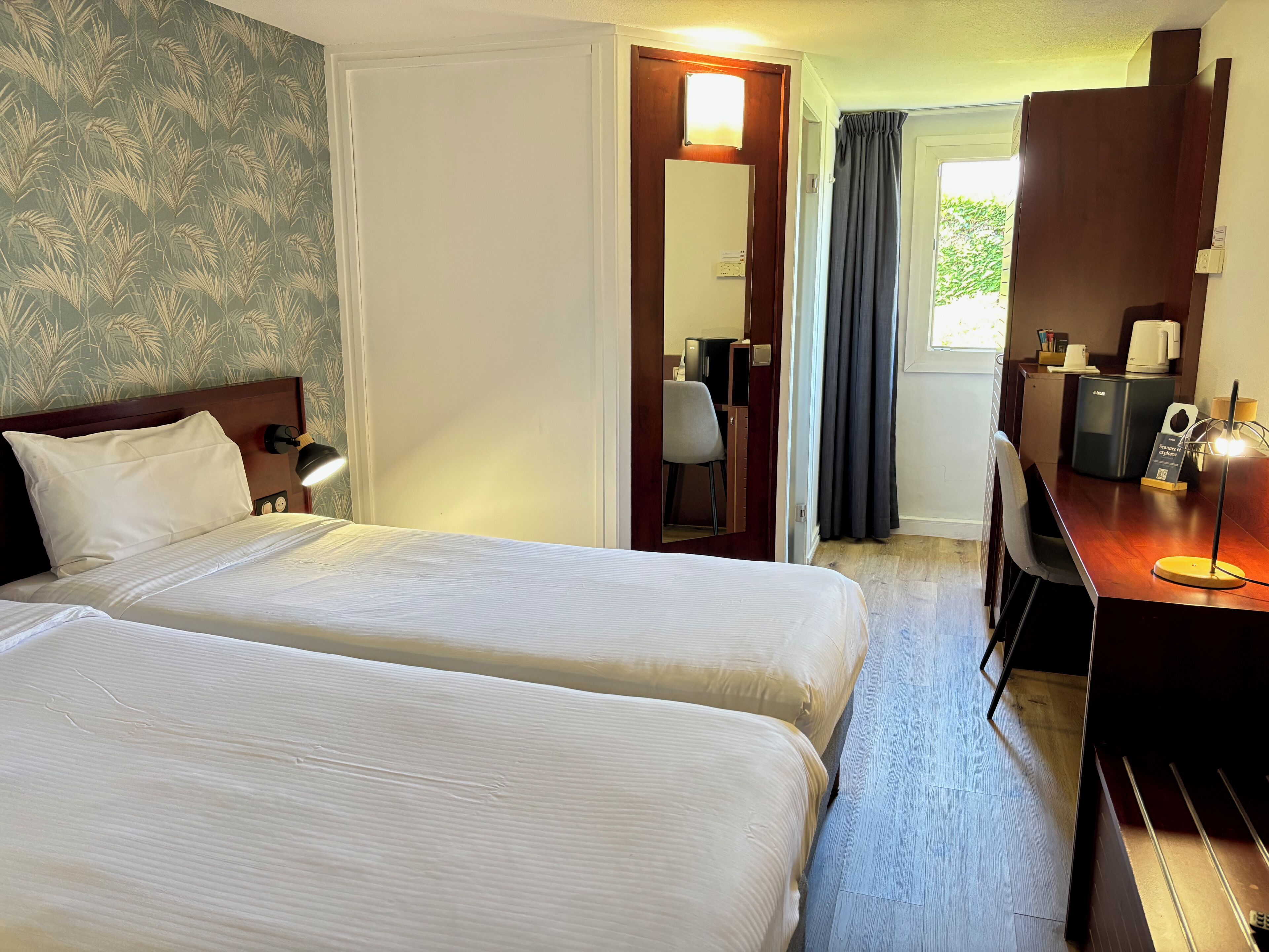 A hotel room with two beds, a desk with a computer, a chair, and a window with curtains.