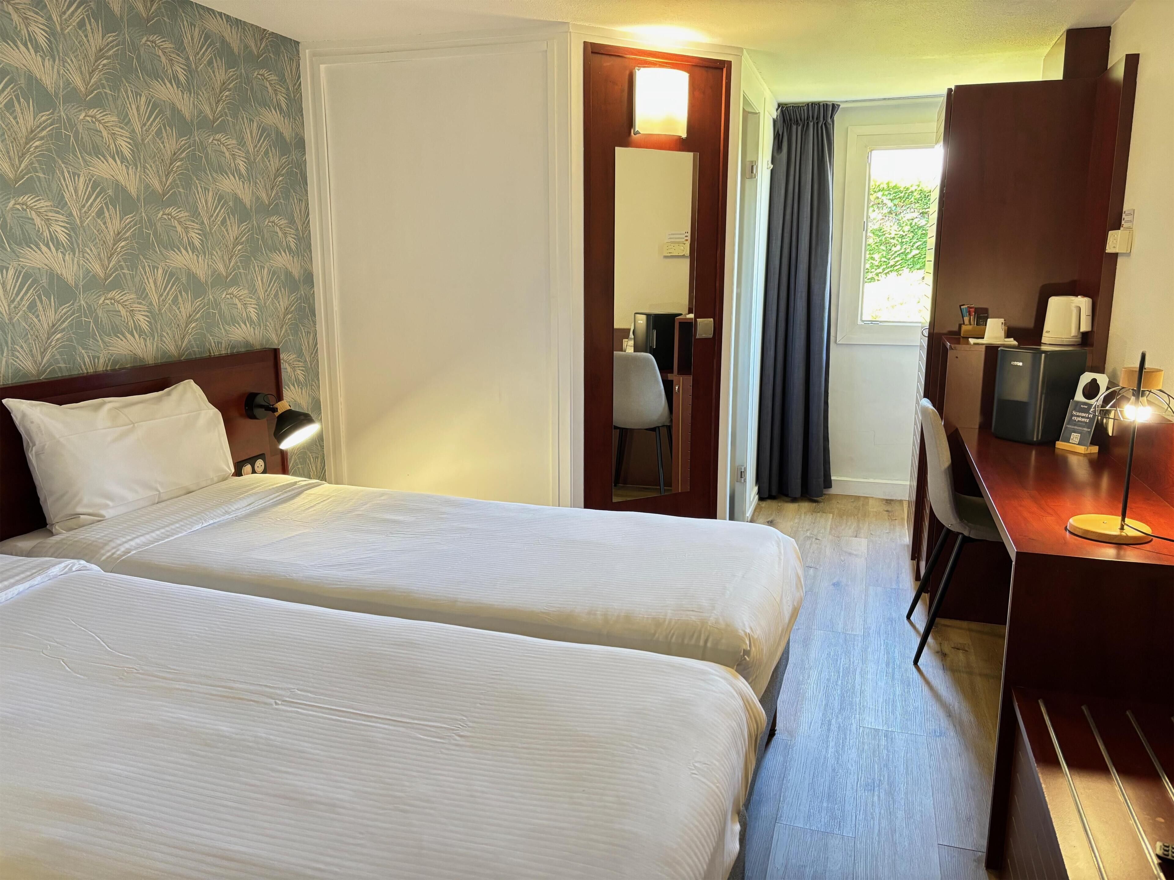 A hotel room with two beds, a desk, a chair, a mirror, and a window with curtains.