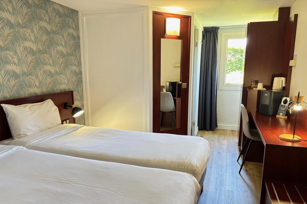 A hotel room with two beds, a desk, a chair, a mirror, and a window with curtains.