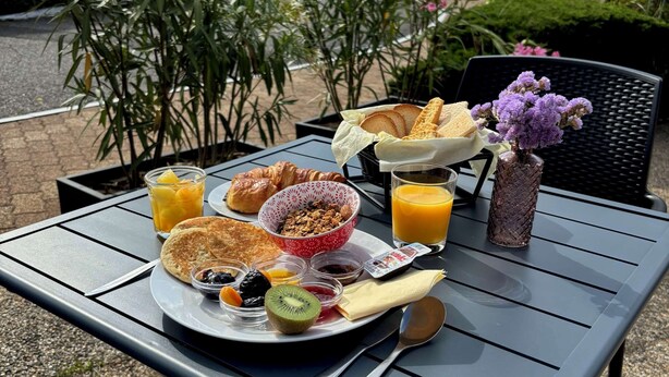 Une table de petit-déjeuner avec des croissants, un panier de fruits, un verre de jus d’orange et un vase de fleurs.