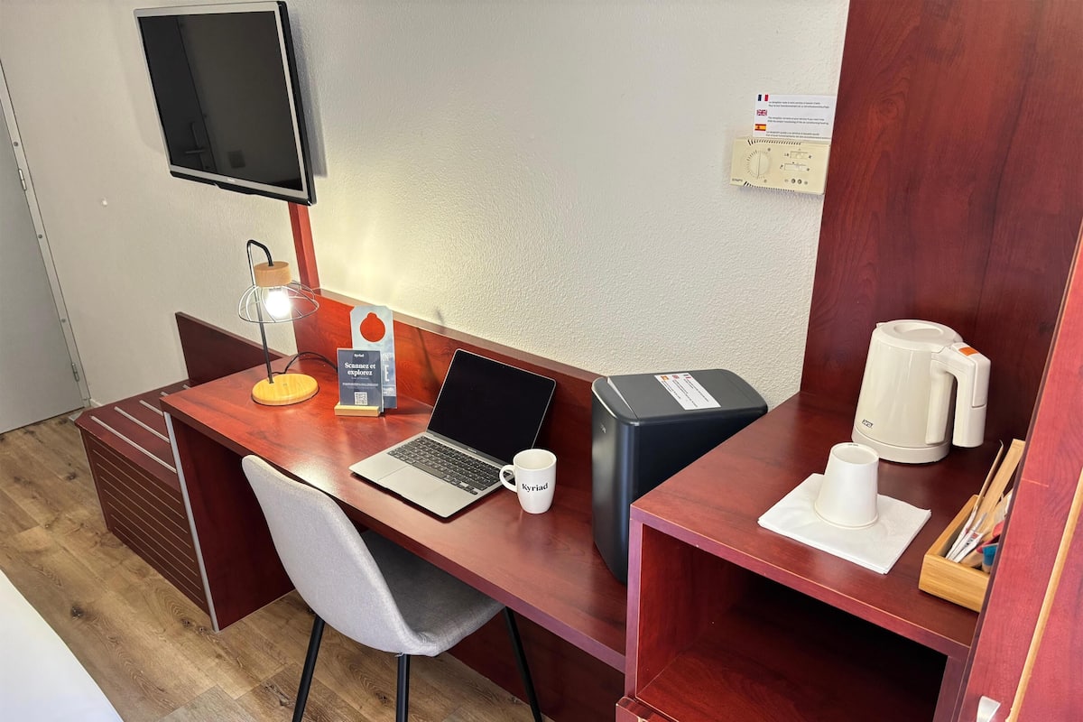 Quarto de hotel com uma escrivaninha de madeira, um laptop, uma máquina de café, uma caneca e uma televisão fixada na parede.
