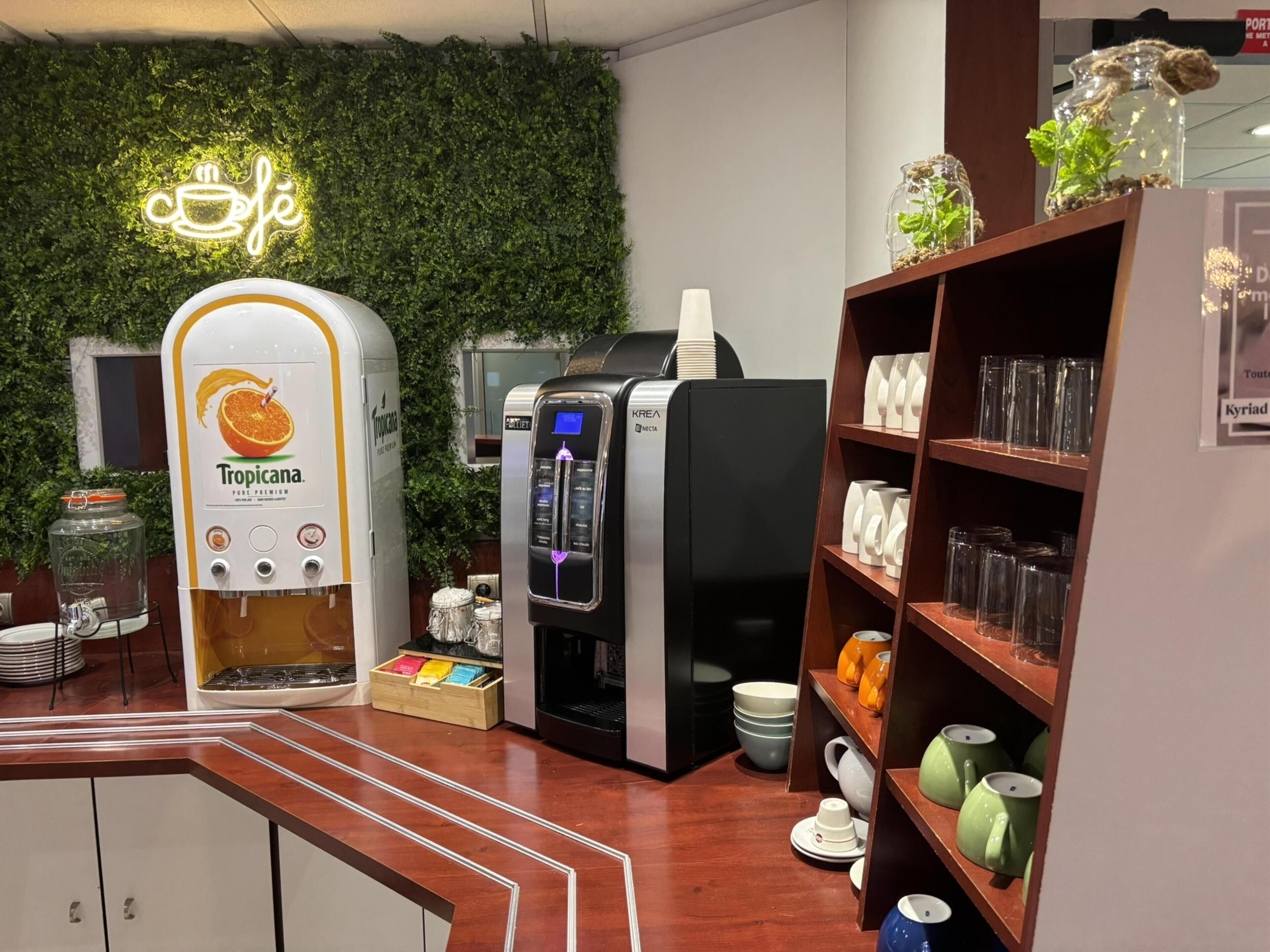 Una estació de cafè amb un dispensador Tropicana, una màquina de cafè KREA i una prestatgeria amb tasses i vasos.