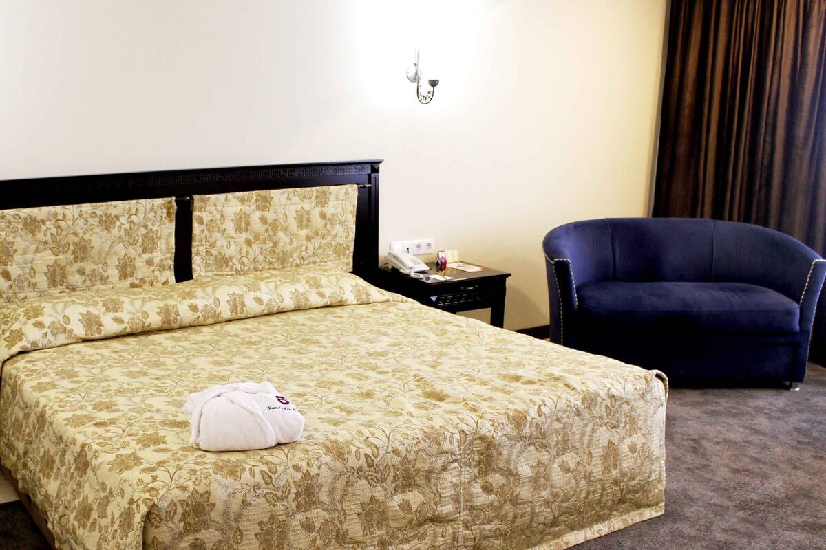 A neatly made bed with a floral bedspread, a bedside table with a phone, and a dark blue armchair.
