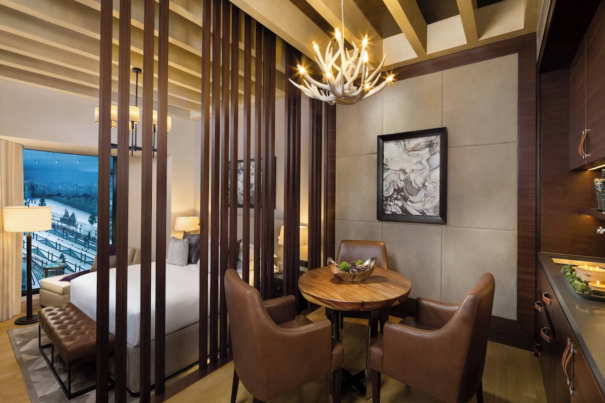Une chambre d’hôtel avec un lit, une table à manger et des chaises, un lustre, et une vue sur un paysage enneigé.