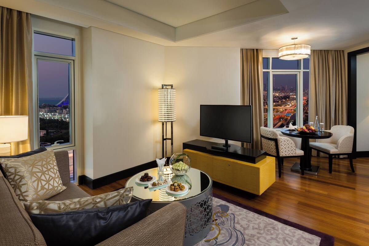 Une chambre d’hôtel moderne, dotée d’un canapé, d’une télévision, d’une table à manger avec des chaises, et offrant une vue sur le paysage urbain la nuit.