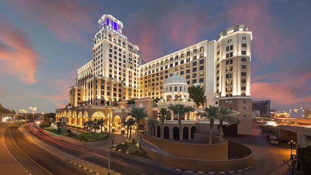 Un hotel grande con varios pisos, una torre destacada con señalización iluminada y un área de entrada bien iluminada con palmeras.