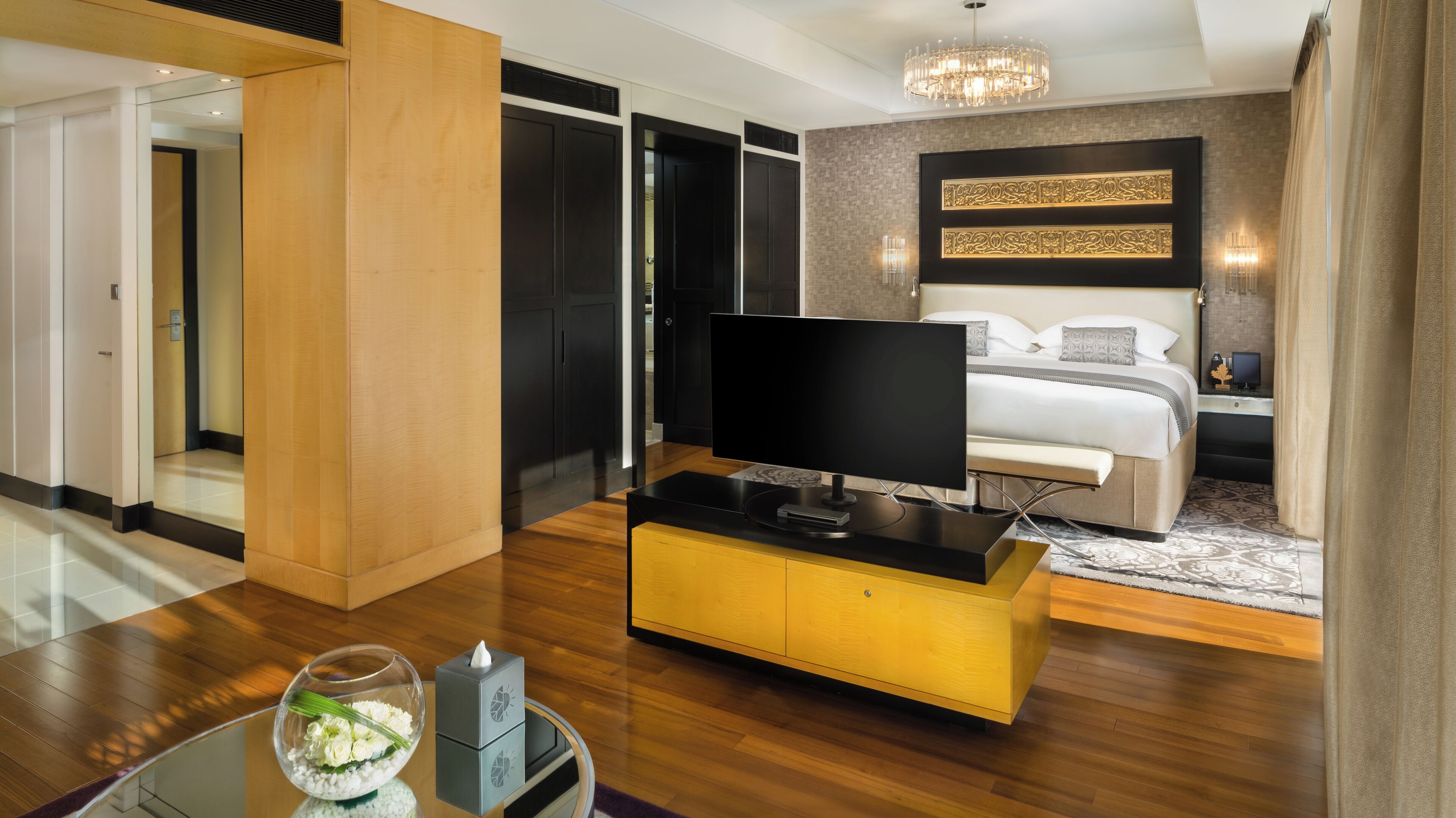 Ein modernes Hotelzimmer mit Flachbildfernseher auf einer Holzkonsole, einem Bett mit weißer Bettwäsche, einem großen Spiegel und einer Chandelier.