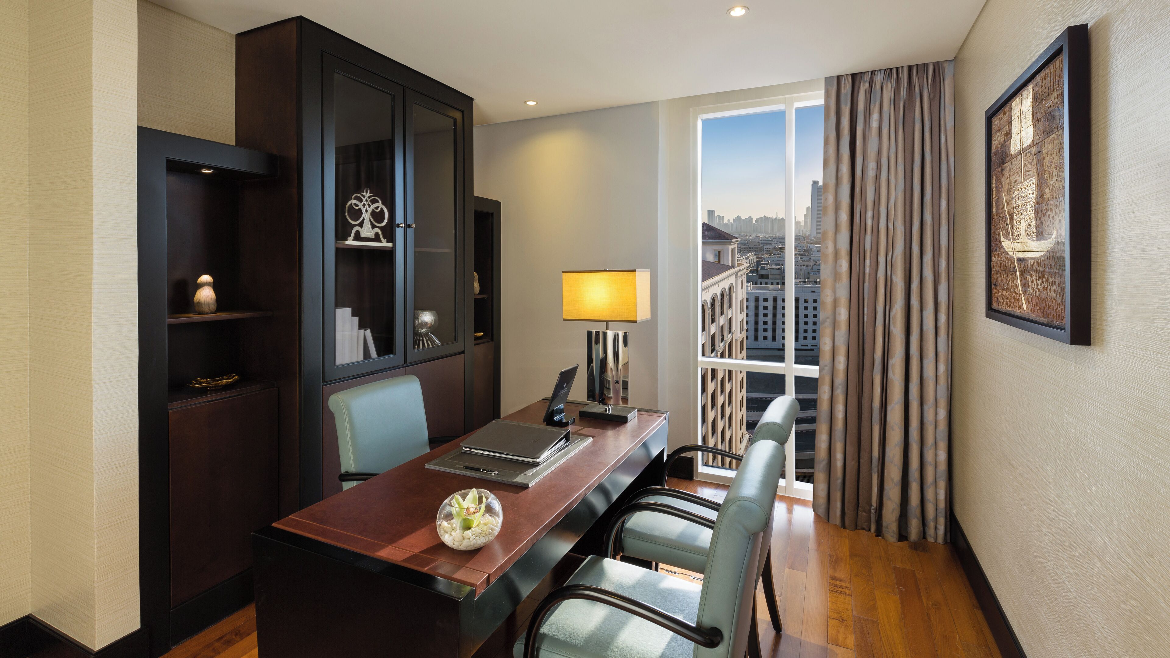 A hotel room with a dining table, chairs, a lamp, a cabinet, and a large window with a city view.
