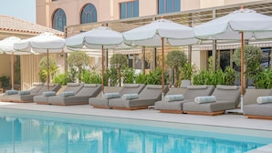Un área junto a la piscina con sillones de descanso y sombrillas, una piscina cristalina y un edificio con ventanas arqueadas al fondo.