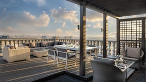 Una terraza en la azotea con vistas al horizonte de la ciudad, amueblada con sofás de mimbre, una mesa con cubierta de vidrio y una mesa de comedor para dos.
