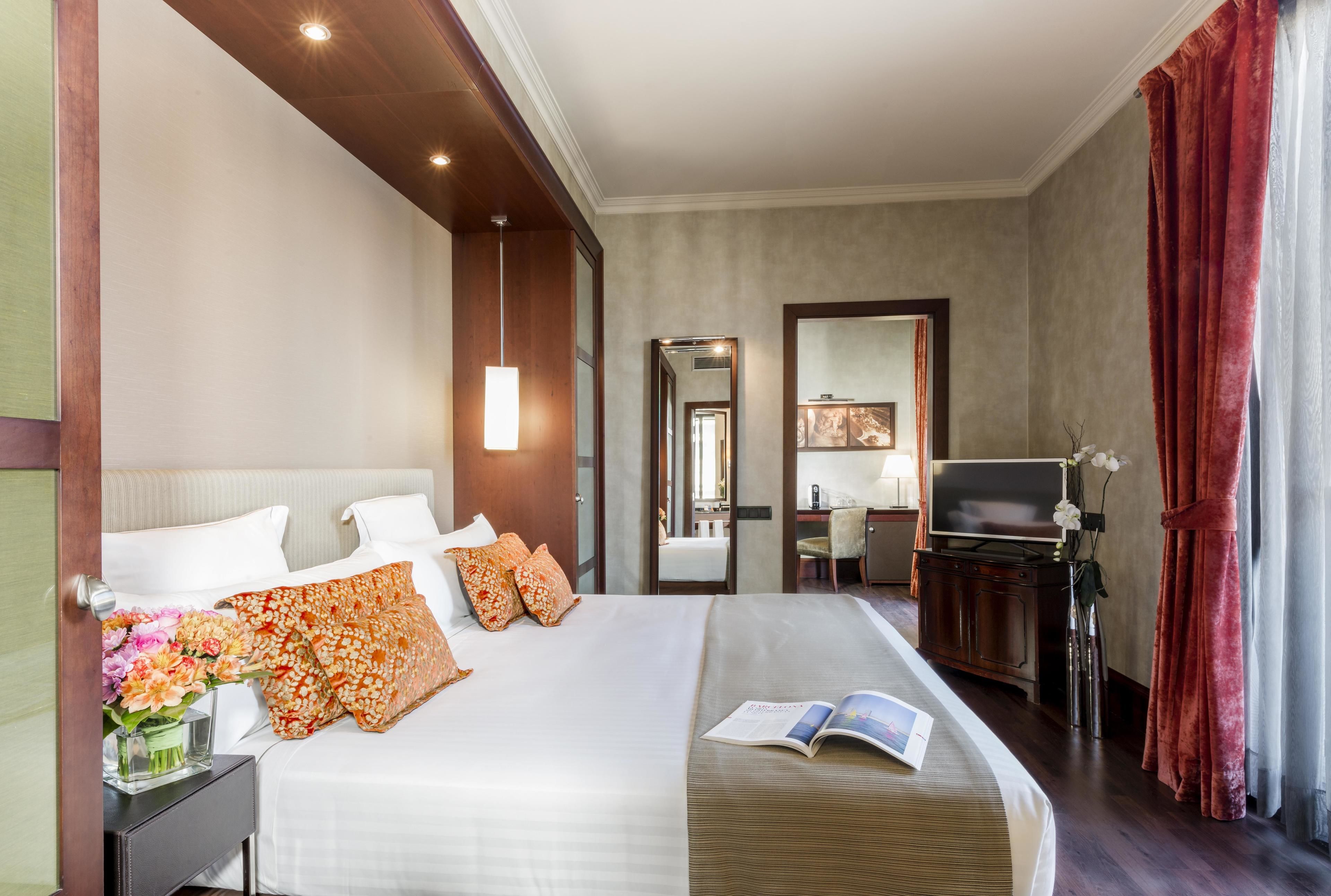 A hotel room with a large bed, a desk with a TV, a mirror, and a window with curtains.