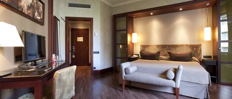 Quarto de hotel com cama, mesas de cabeceira, escrivaninha com cadeira, televisão e obras de arte na parede.