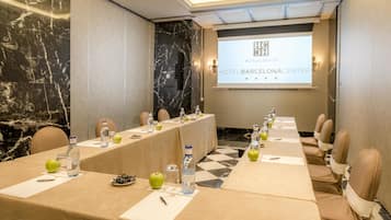 Een congrescentrum met een lange tafel, stoelen, waterflessen, appels en een scherm waarop 'HOTEL BARCELONACENTER' staat.