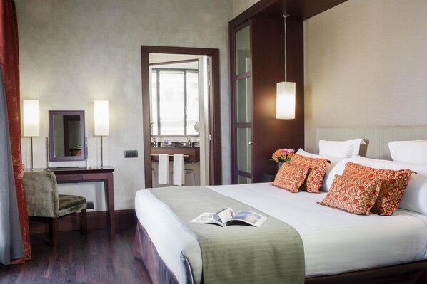 Habitación de hotel con cama, escritorio, silla, espejo y baño visible a través de una puerta abierta.