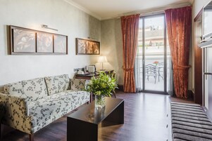 A living room with a floral sofa, a coffee table with a vase of flowers, a lamp, and a sliding glass door leading to an outdoor area with a table and chairs.