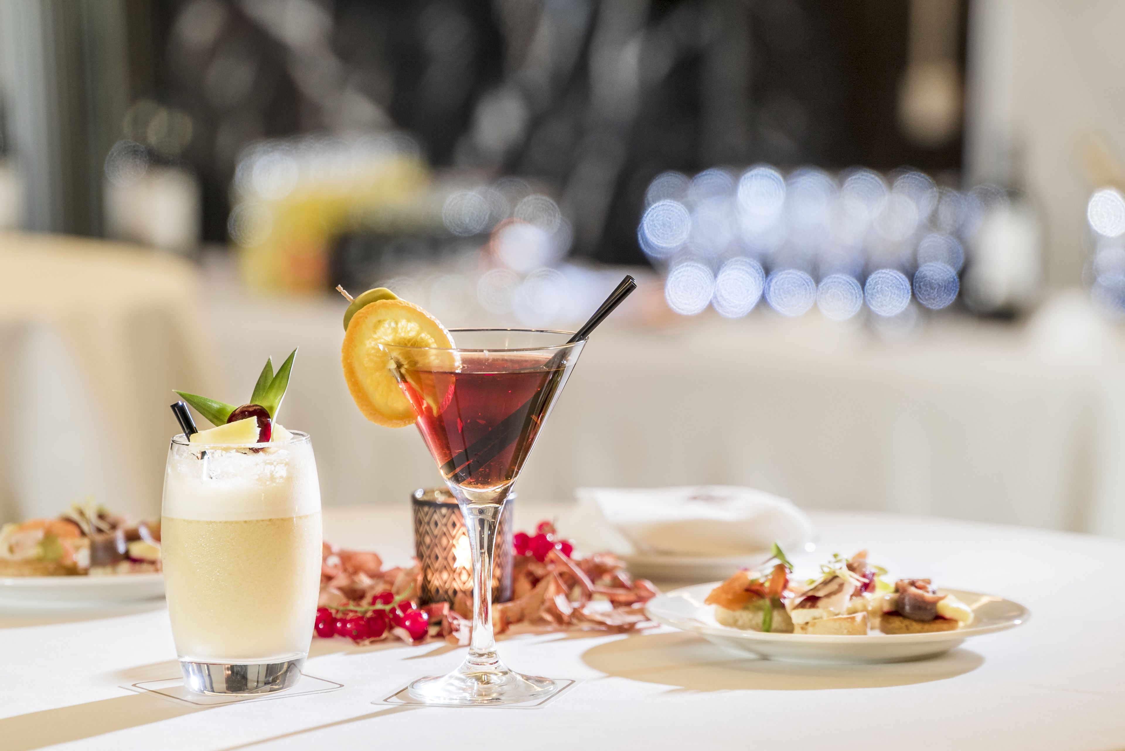 A table with a martini glass and a highball glass, both with garnishes, and a plate of food in the background.