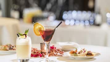 Een tafel met een martini-glas en een highball-glas, beide met garnering, en een bord eten op de achtergrond.