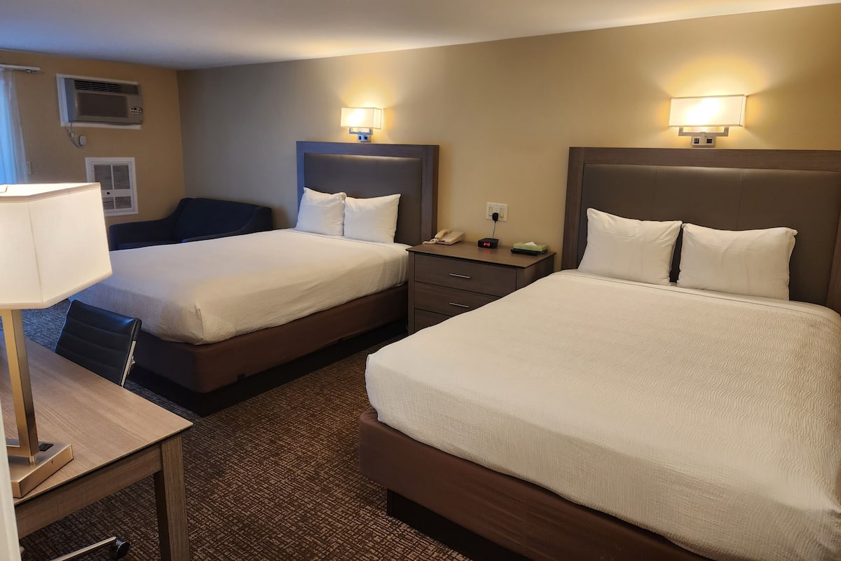 A hotel room with two beds, a desk, a chair, a lamp, and a nightstand with a phone and a small table.