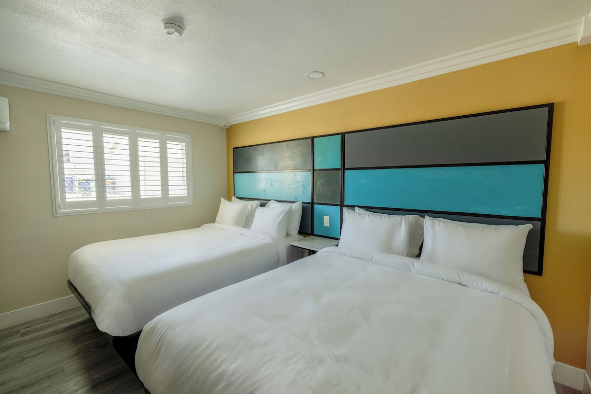 A hotel room with two single beds, a window with blinds, and a wall with a geometric design.