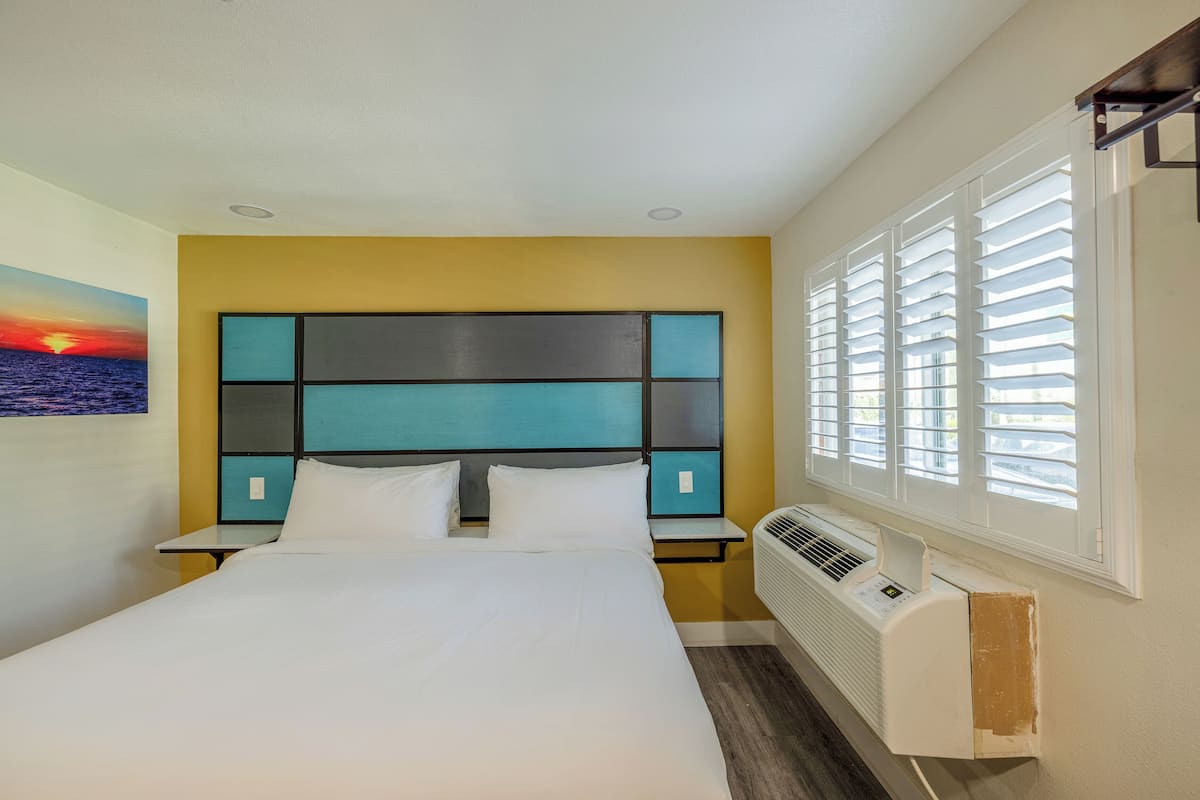 A neatly made bed in a hotel room with a modern headboard, a nightstand, and a window with blinds.