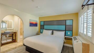 Habitación de hotel con cama, baño con lavamanos y espejo, un cuadro de un atardecer y una ventana con persianas.