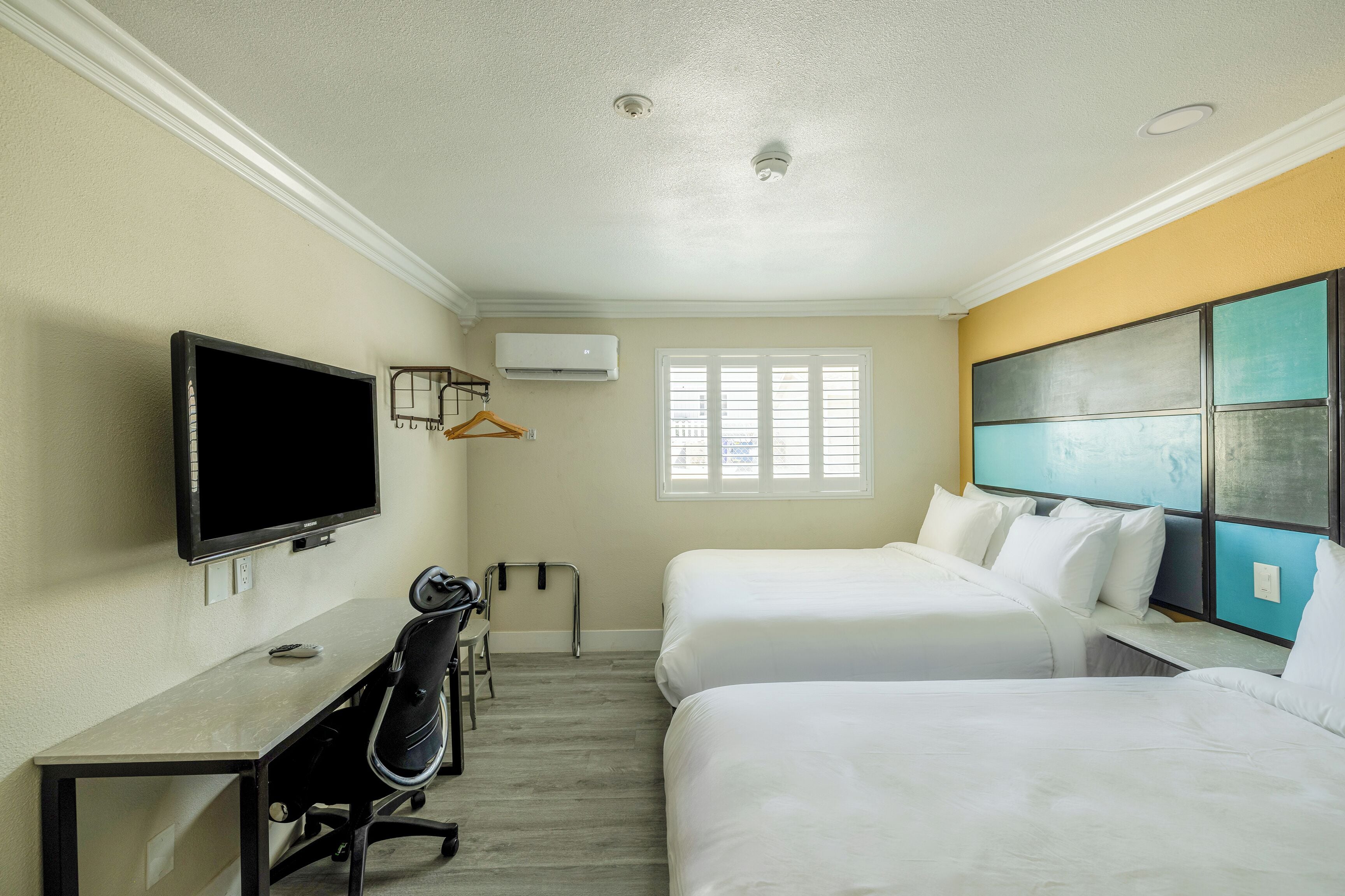 A hotel room with two beds, a desk, a chair, a TV, and an air conditioning unit.