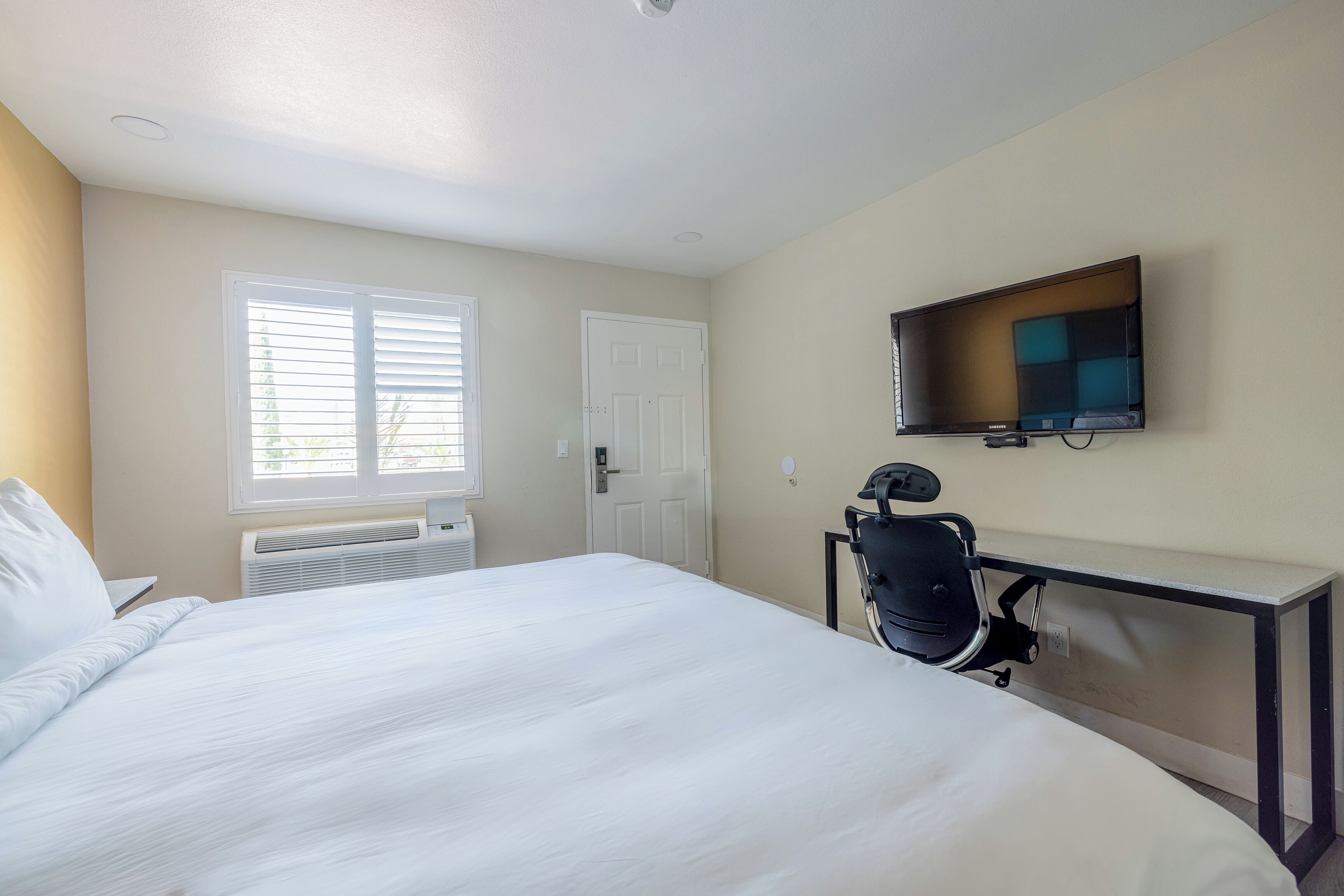 A hotel room with a bed, a desk, a chair, a TV, and a window with blinds.
