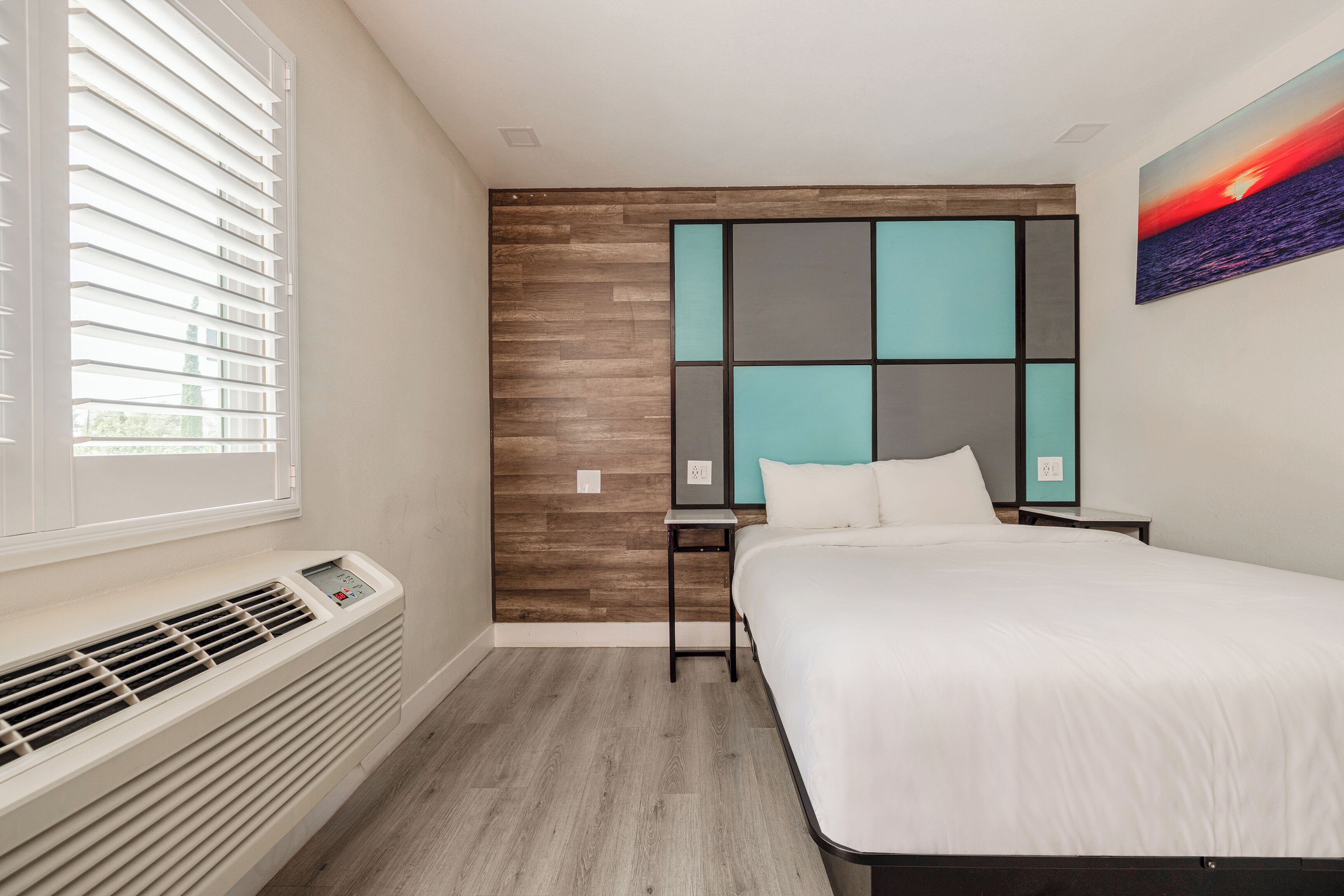 A modern hotel room with a large bed, a built-in heater, a window with blinds, a wall-mounted artwork, and a small table.