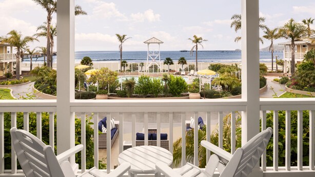 Un balcón con sillas y una mesa blancos, con vistas a una zona de piscina con palmeras y al océano.