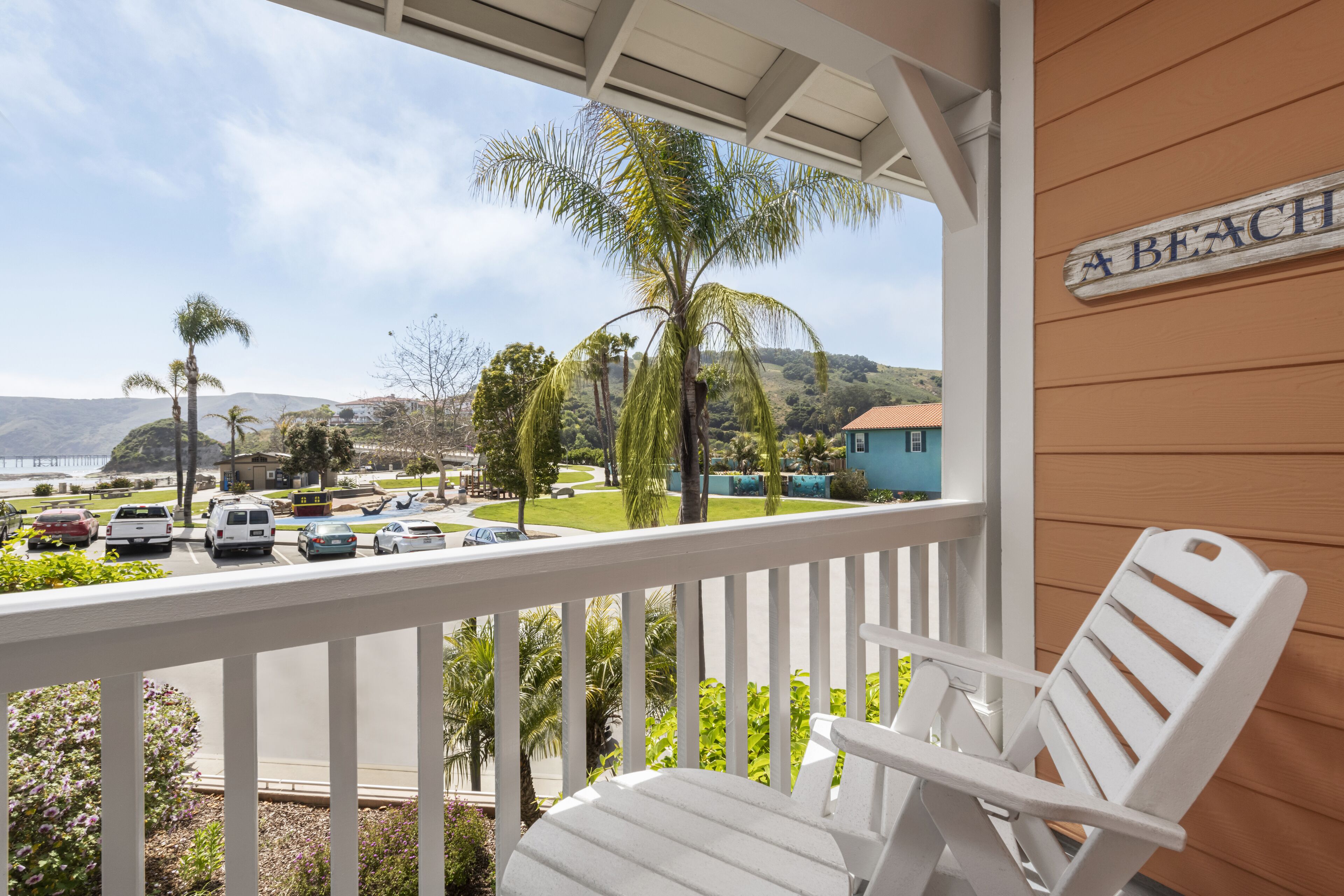 Un balcon avec une chaise blanche, une pancarte indiquant « UNE PLAGE », et une vue sur un parking et des espaces verts.