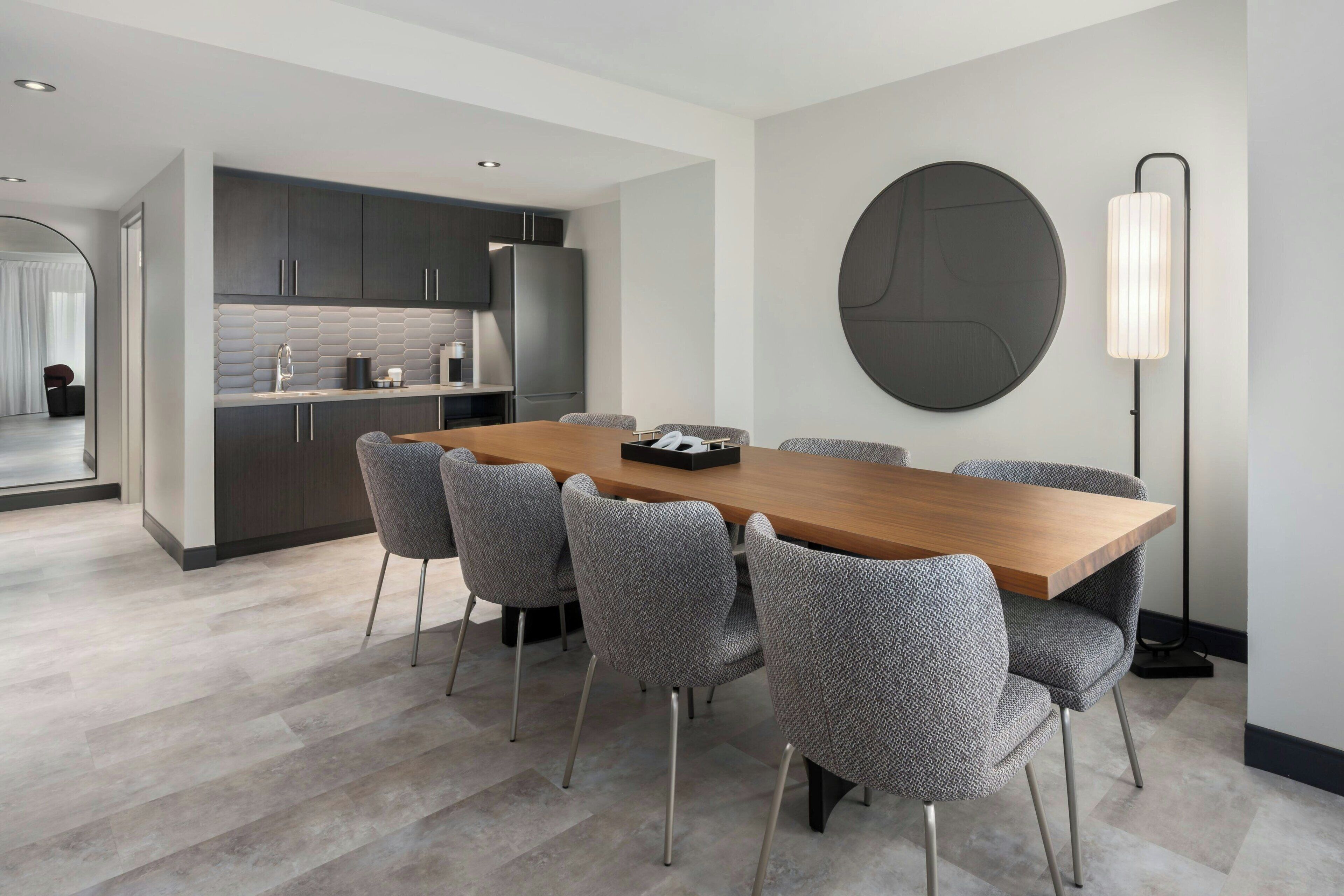 Een moderne eetruimte met een houten tafel en zes grijs beklede stoelen. Op de muur hangt een groot zwart rond kunstwerk, en rechts is een vloerlamp zichtbaar.